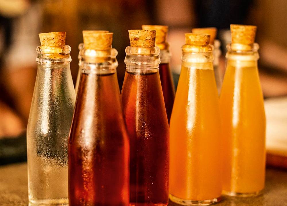 Sete drinques engarrafados de diferentes cores em vasilhames pequenos tampados por rolha sobre bancada do Cava Bar.