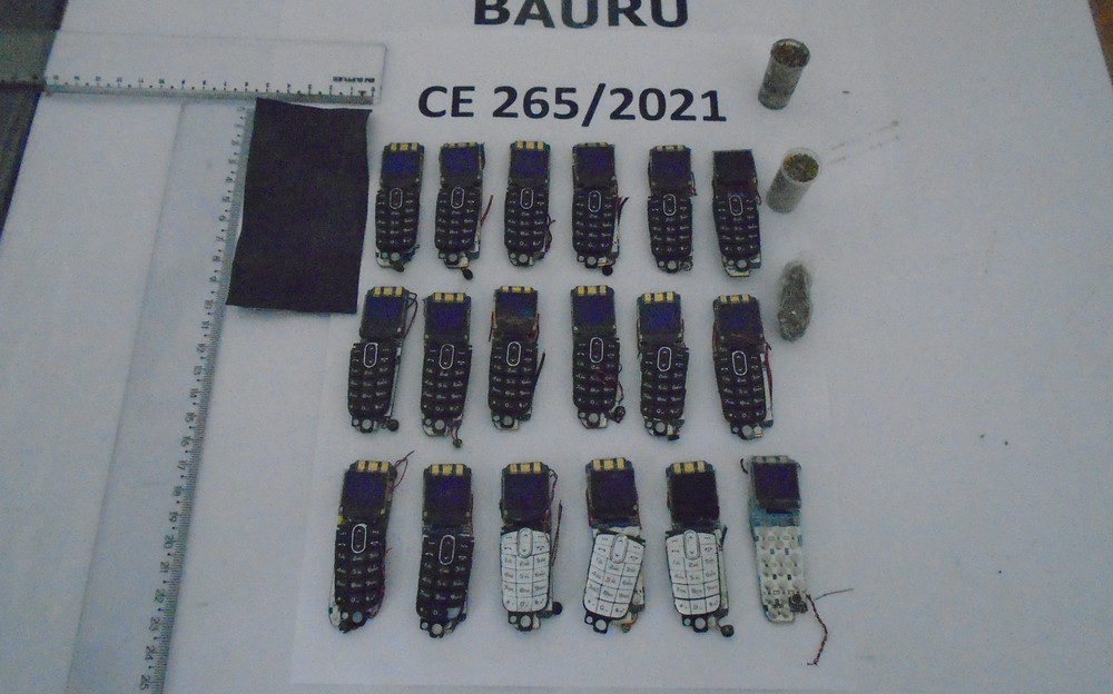 Celulares Detentos corpo A imagem mostra 18 celulares deformados em cima de uma mesa branca com réguas ao lado