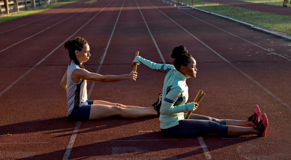A imagem mostra duas atletas no longa sentadas numa pista, praticando a troca de bastão
