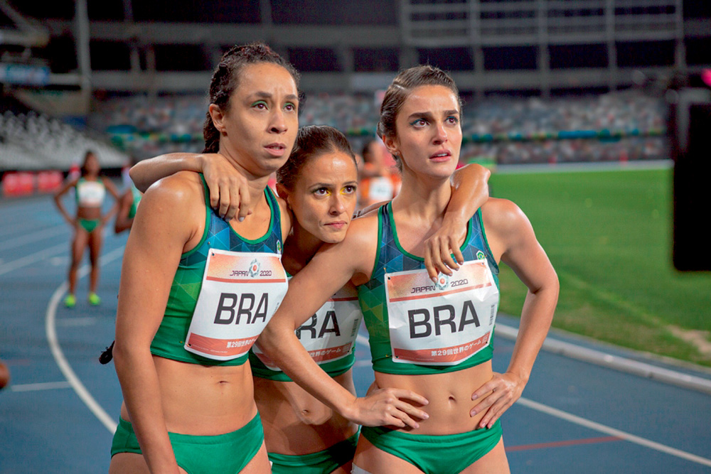 A imagem mostra as três personagens do filme com uniforme do atletismo, abraçadas em uma pista, ansiosas e olhando para cima
