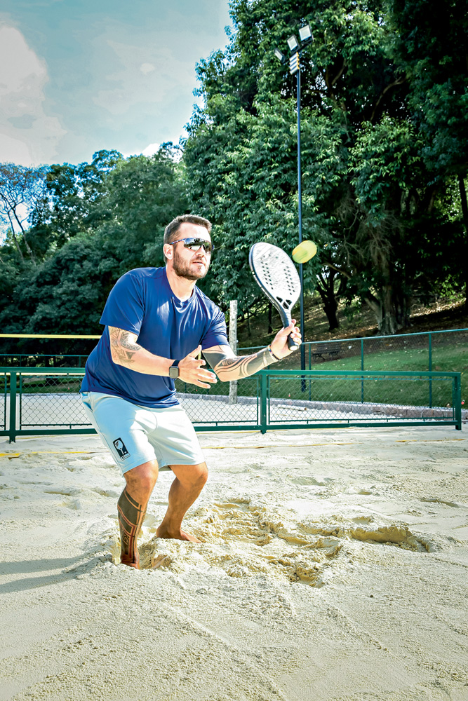 A imagem mostra o professor acertando a bolinha em uma altura média. Ele está em uma quadra de beach tennis, com os joelhos flexionados para acertar a bola na altura do seu ombro.