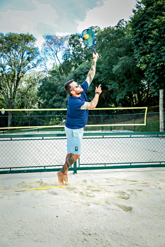 A imagem mostra o professor acertando a bola de beach tennis em uma quadra de areia.