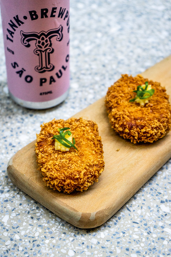 Croqueta – Tank Brewpub Duas croquetas de milho sobre tábua de madeira. Ao lado, latinha com rótulo de cor rosa, com o logo do Tank.