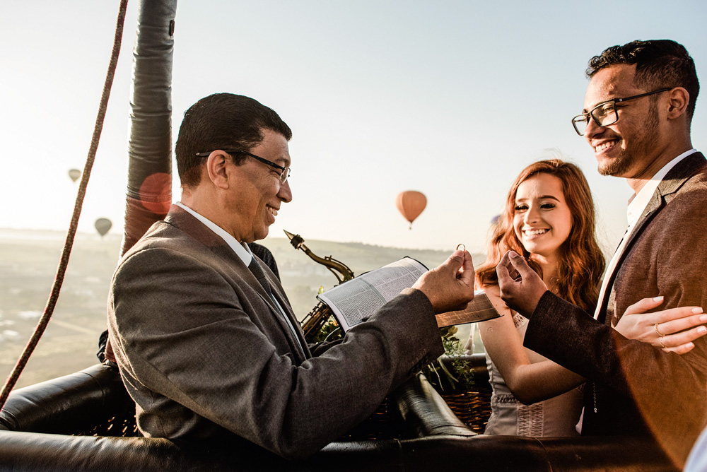 Foto mostra três pessoas em um balão sorrindo. Um homem de terno lê uma bíblia com uma aliança na mão. Do outro lado, o casal se abraça.