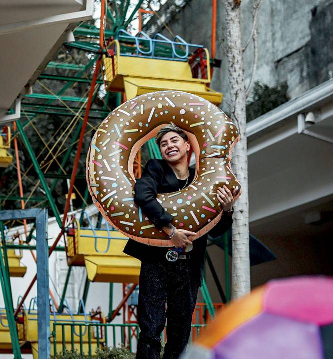Felipe Didonih abraça boia inflável em formato de donut marrom. Veste roupas pretas e aparece de pé em frente a uma roda-gigante instalada em seu quintal.