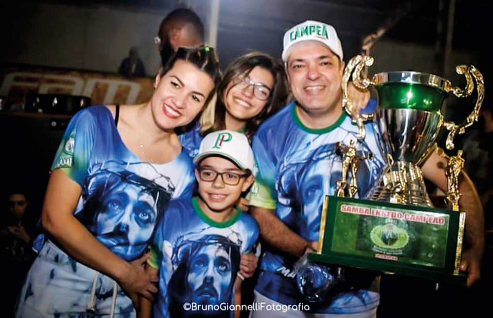 anna e marcelo posando para a foto com os filhos giovanna e guilherme, no carnaval em 2020. Todos estão com camisa da escola de samba Mancha Verde e marcelo está segurando o troféu de campeão do carnaval