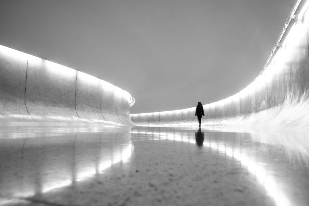 Fotografia em preto e branco mostra longo corredor curvado com iluminação nas extremidades e silhueta de uma pessoa ao fundo.