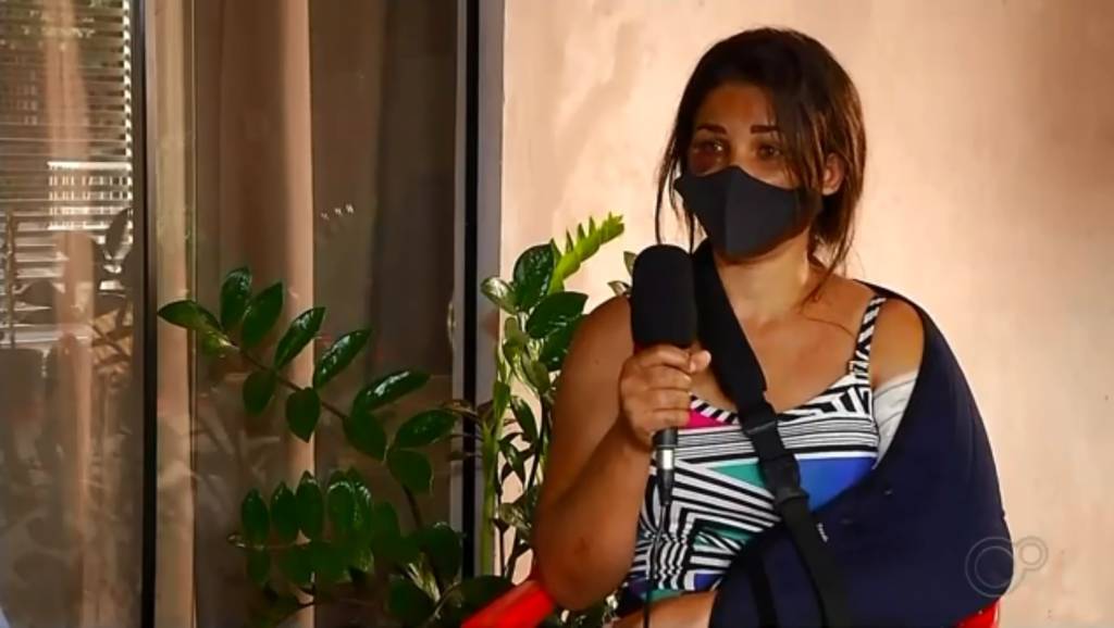 A imagem mostra Adriana, segurando o microfone com a mãe esquerda e o braço direito apoiado em uma tipoia. Sua olho direito está roxo e ela está usando máscara.