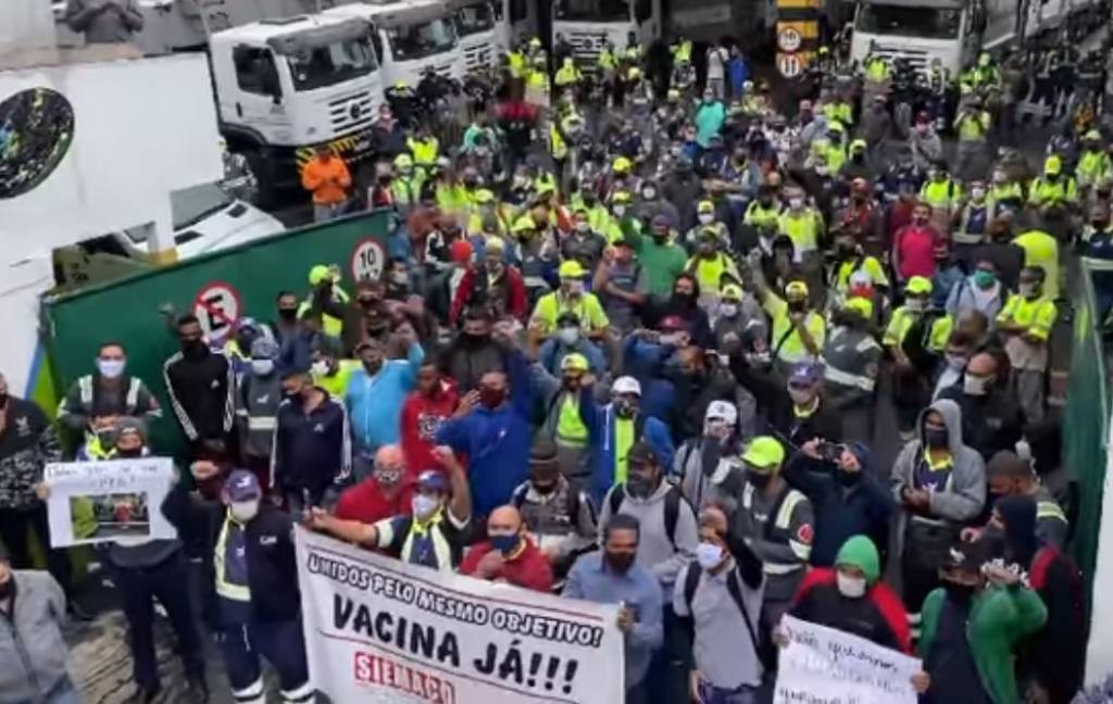 A imagem mostra trabalhadores da limpeza na rua, de uniforme e máscaras, com faixas escritas "Vacina Já!"