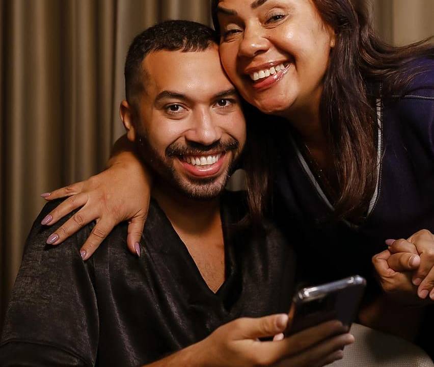A imagem mostra Gilberto sentado sendo abraçado por sua mãe. Ambos estão sorrindo para a câmera.