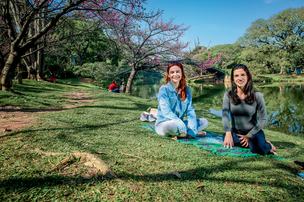 A imagem mostra as duas amigas sentada sobre a grama do Ibirapuera, sorrindo para a câmera.