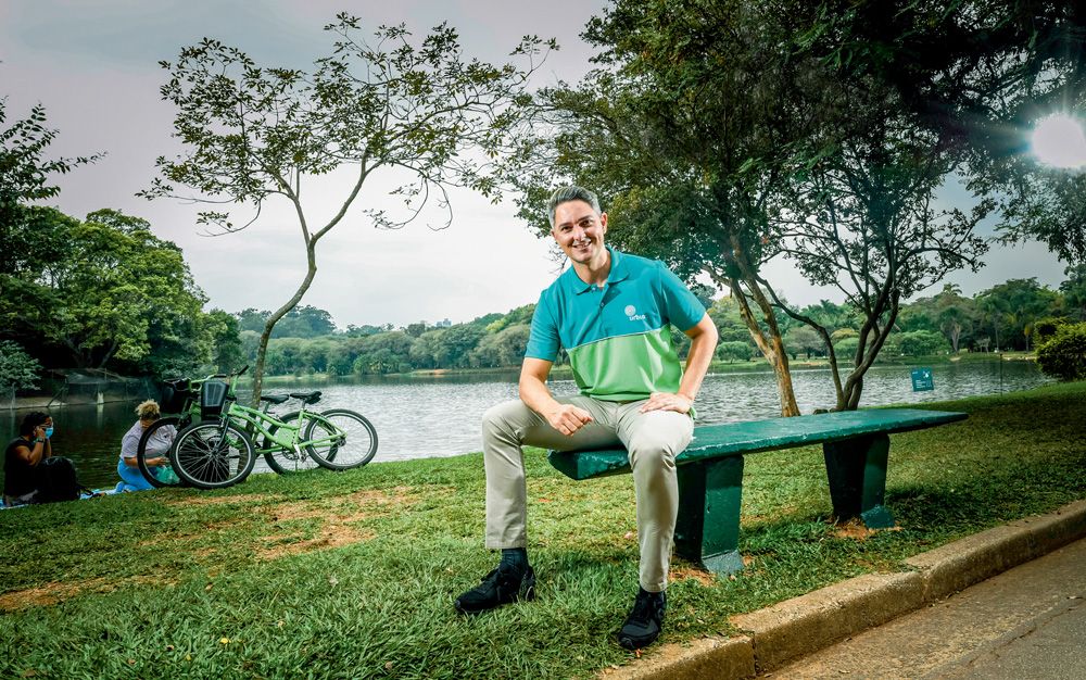 A imagem mostra Lloyd sentado em um banco no parque Ibirapuera. Ele sorri para a câmera com a mão no seu joelho.