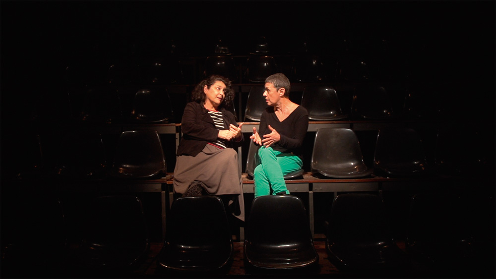 Duas mulheres conversam em uma plateia de teatro escura