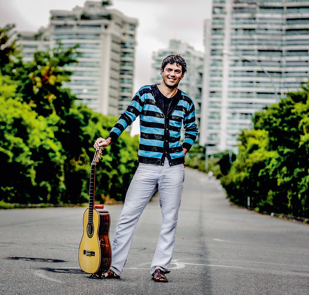 Um homem posa com um violão apoiado no chão ao seu lado. Ele está sorrindo em um cenário com prédios e árvores