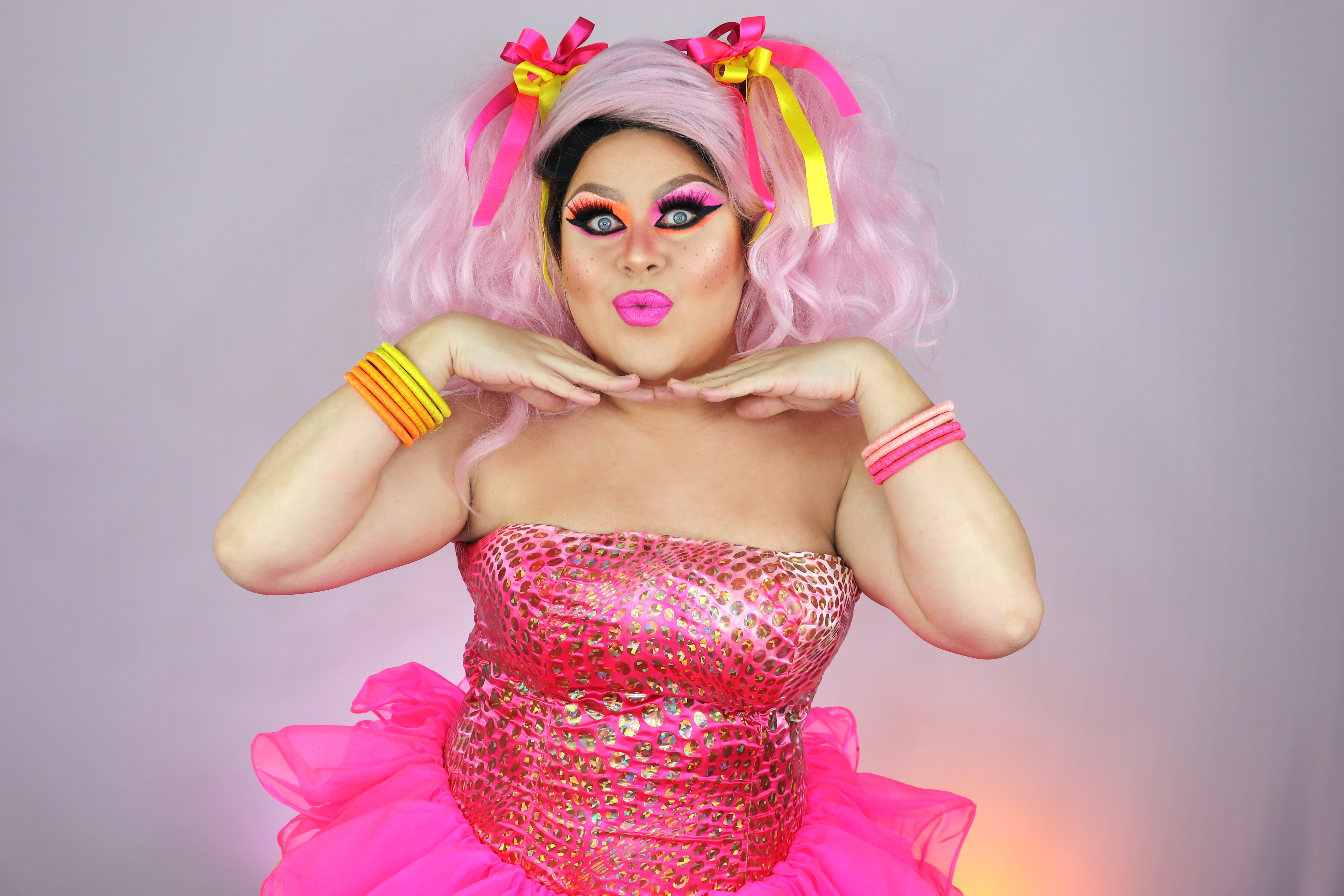 Drag queen posa montada com peruca, vestidos e acessórios rosa. Está com as mãos no queixo e fazendo bico com a boca