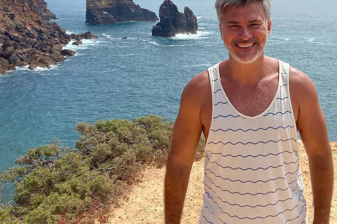 A imagem mostra Leonardo em uma praia, com o mar do fundo, e ele de frente para a câmera, olhando sorrindo para ela e com uma camiseta regata