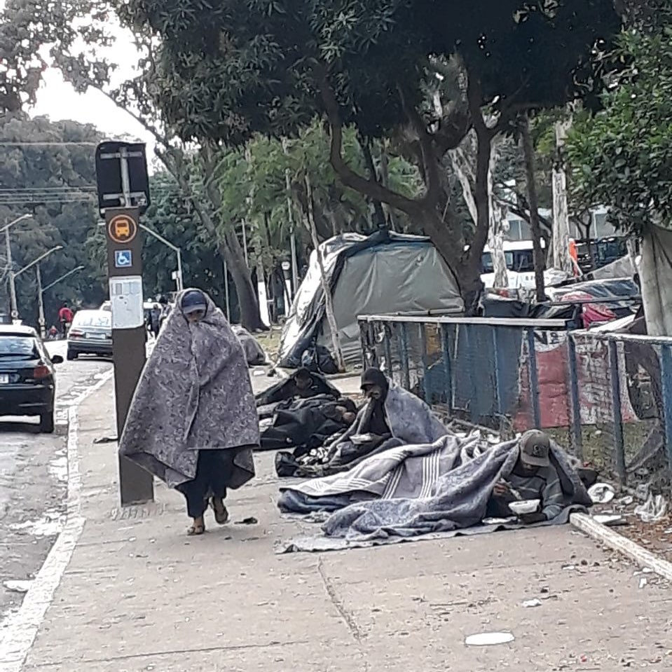 A imagem mostra três moradores de frio com grandes cobertores. Dois estão deitados na rua e um está caminhando.