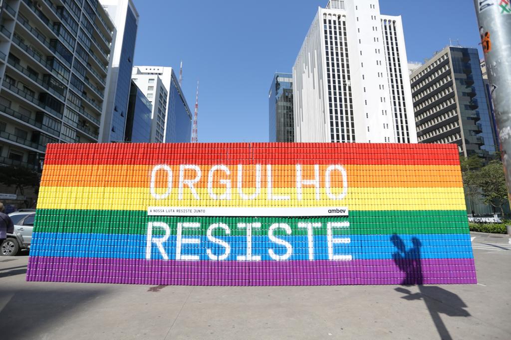 Mural de latas de alumínio forma as cores do arco-íris e a frase "Orgulho Resiste".