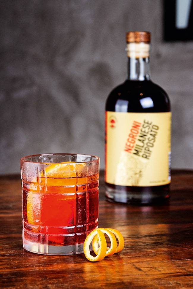 Sobre bancada de madeira, garrafa de Negroni ao fundo à direita e mais a frente um copo com a bebida servida mais fatia de laranja.