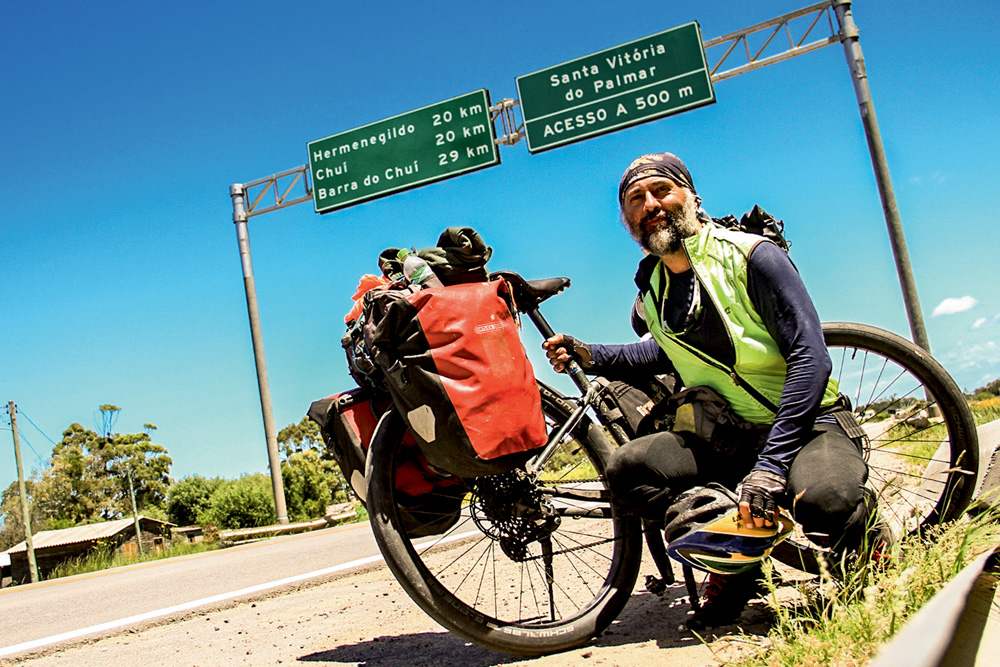 Nestor Freire aparece agachado ao lado de sua bike. Veste colete verde e bandana. Ao fundo, na rodovia, a foto mostra duas placas verdes grandes.