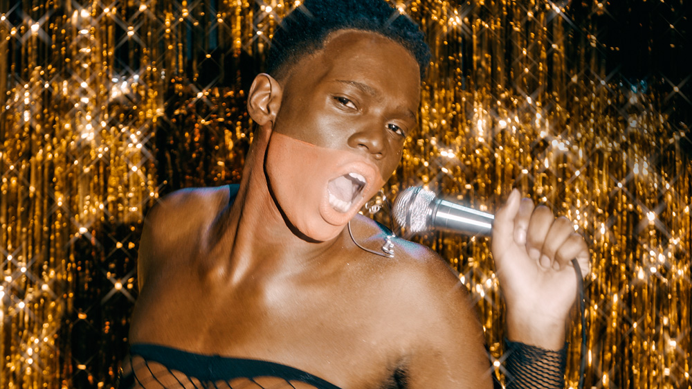 homem negro com detalhe no rosto cantando com uma cortina dourada brilhante no fundo