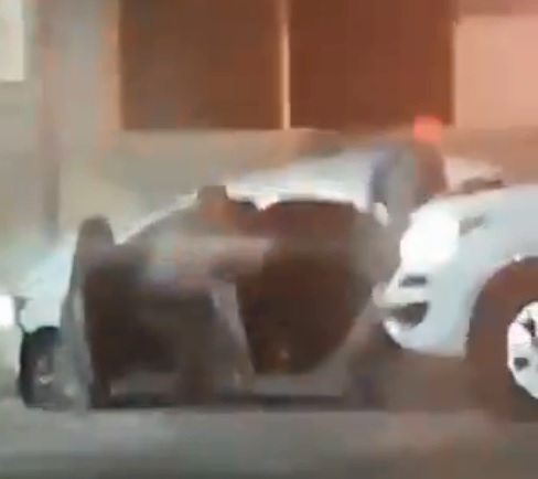 A imagem mostra um policial militar ajoelhado, em frente ao carro branco, apontando a arma pra dentro.