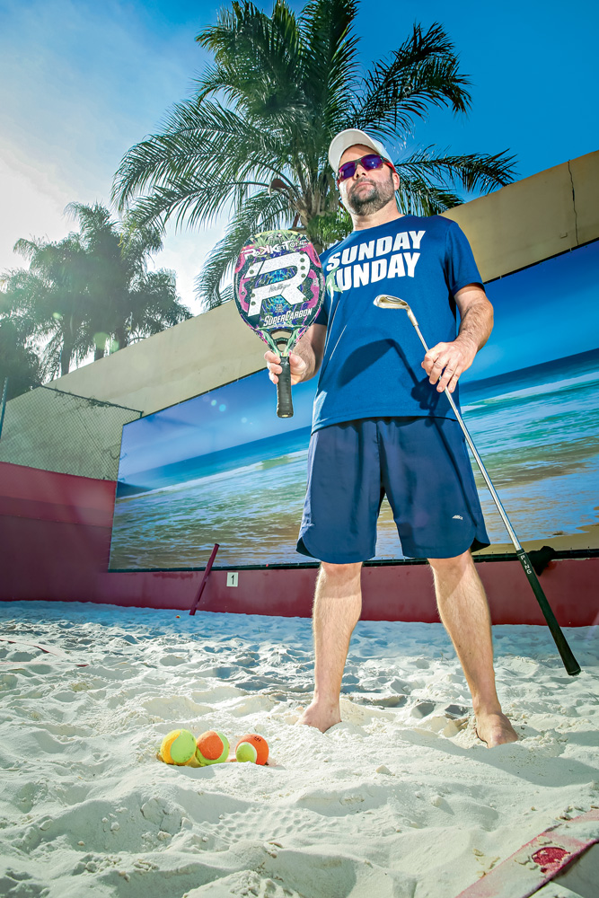 A imagem mostra Philippe, em uma quadra de beach tennis, com sua raquete e taco de golf. Ele está com óculos escuros e boné.