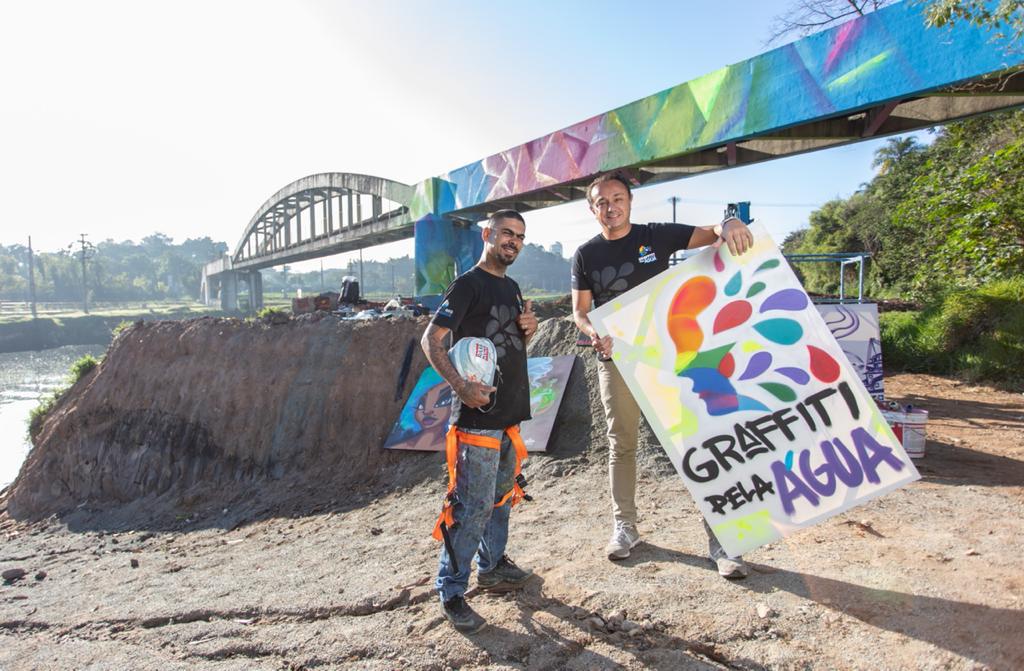 Gamão (esquerda) e Rodrigo Cordeiro, idealizadores do projeto Graffiti pela Água. Ambos posam em frente a ponte colorida. Rodrigo segura placa com título 