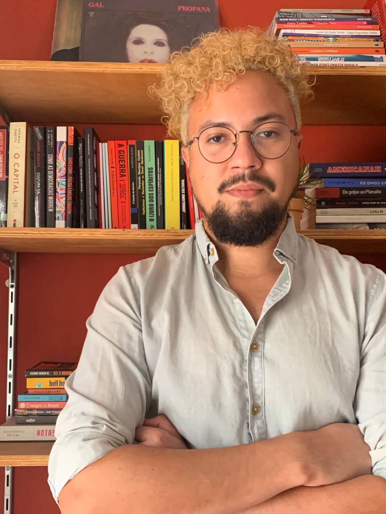 Raul Perez posa de óculos e cabelos encaracolados descoloridos em frente a estante de livros.
