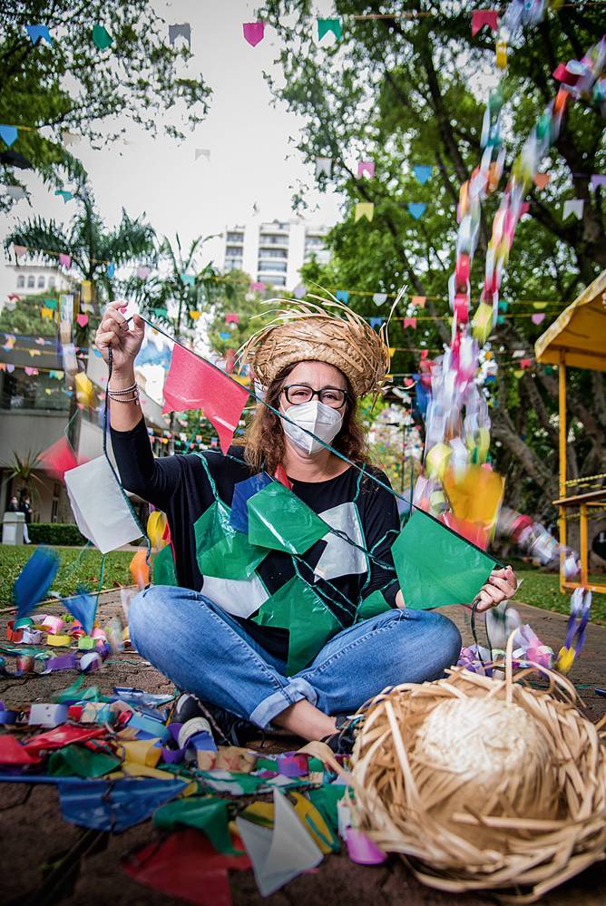 Mulher de máscara sentada com pernas cruzadas mostra bandeirinhas coloridas de festa junina