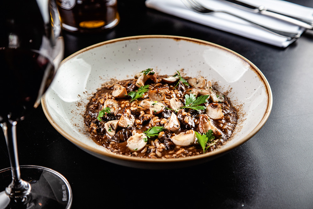 Prato fundo com risoto de cogumelos ao lado de taça de vinho tinto.