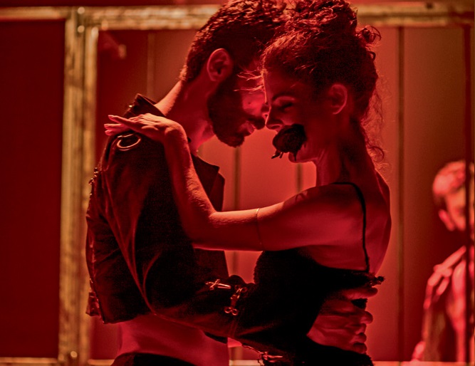 Com luzes bem avermelhadas, foto mostra um homem e uma mulher dançando juntos. Ela tem uma rosa na boca