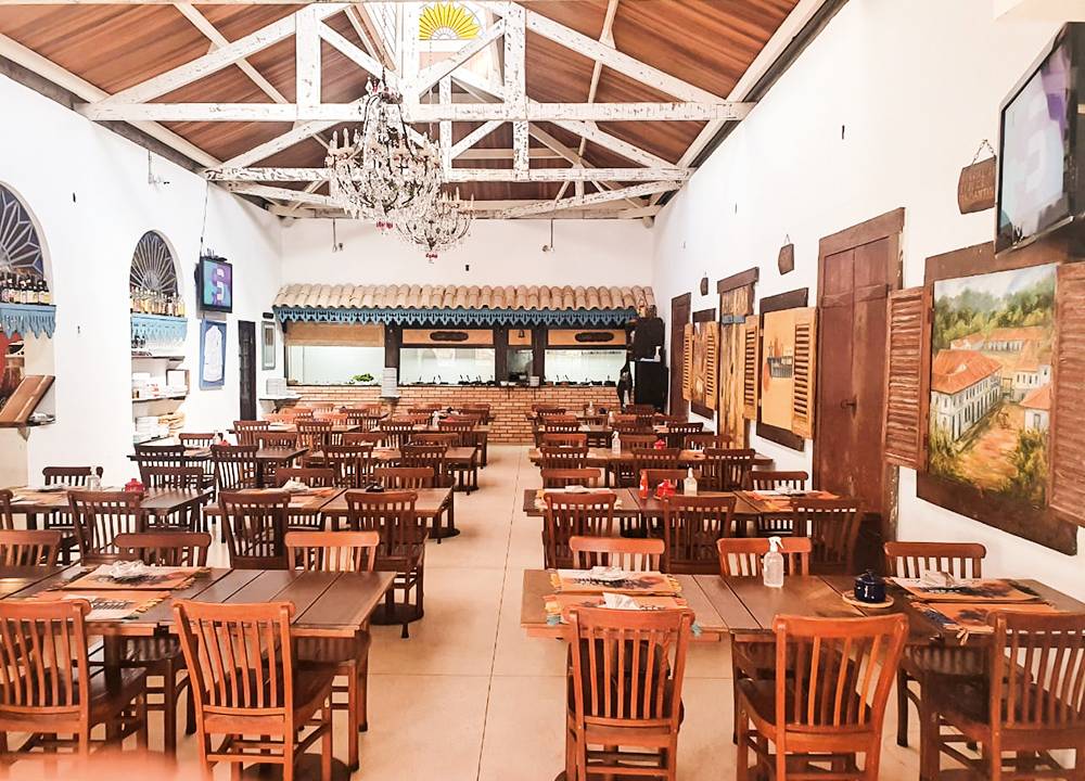 Salão amplo do restaurante Dona Lucinha, com pé direito alto, paredes pintadas de branco, com quadros pendurados. Mesas de madeira espalhadas pelo salão.