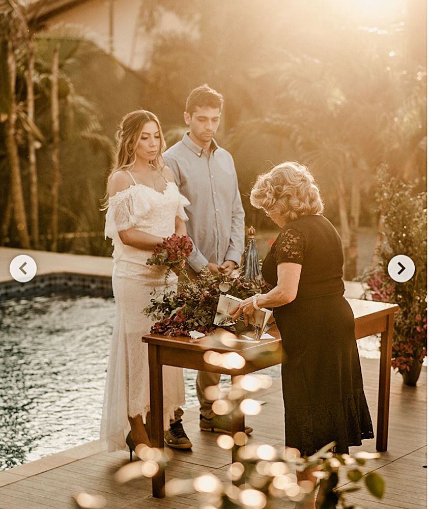 Em frente a uma piscina, dois noivos, um homem e uma mulher, estão em frente a uma senhora cerimonialista. Uma mesa com decorações separa eles