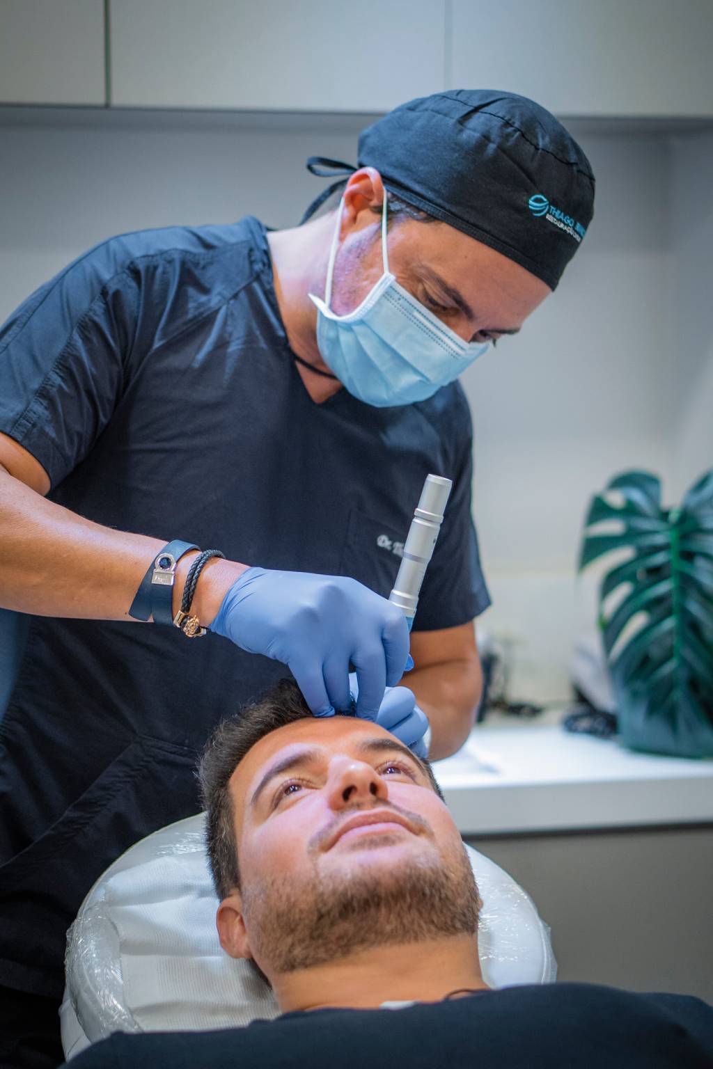 Doutor Thiago Bianco aplica agulha na cabeça de um paciente durante transplante capilar. Usa máscara protetora e veste roupa cirúrgica na cor azul.