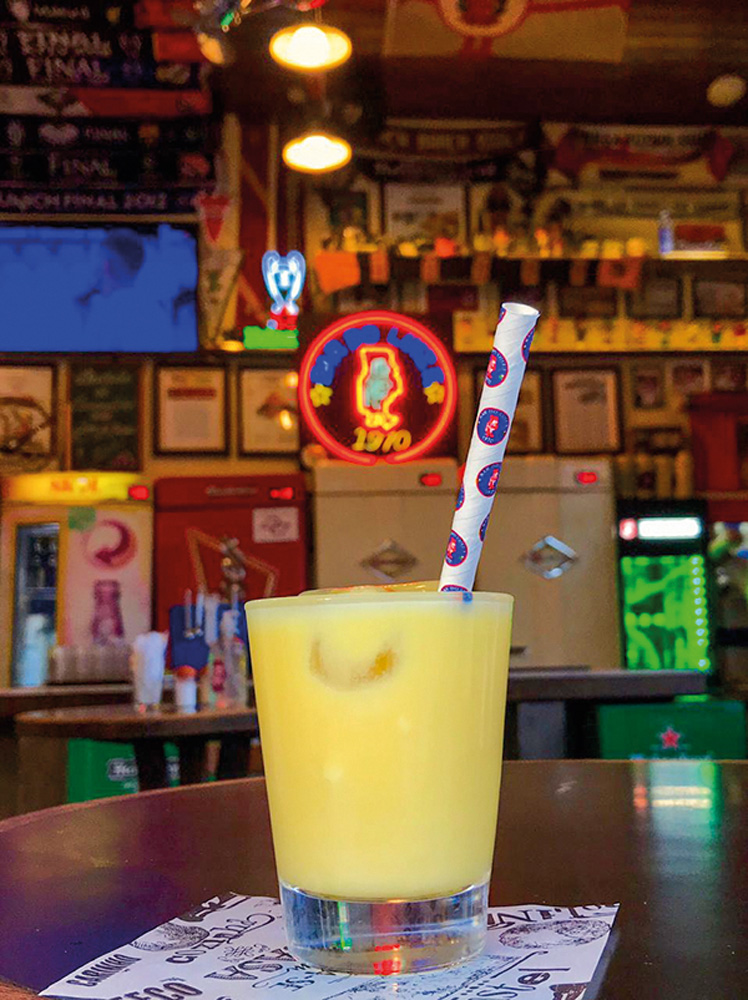 Batida amarela de curau em copo de vidro com canudo colorido sobre mesa de bar.