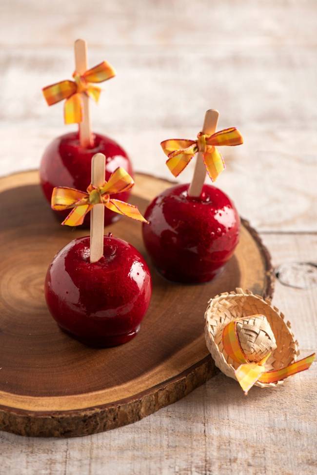 Três maçãs do amor (com caramelo vermelho por fora) postadas de cabeça para baixo com palito de madeira adornada por lacinho laranja sobre tábua redonda de madeira.