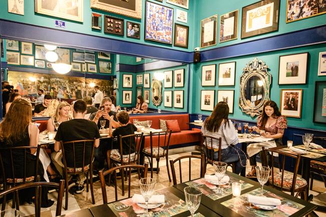 Ça-Va-Erick-Jacquin Salão do restaurante Ça-Va. Com paredes verde-água cobertas de quadrinhos e mesas com pessoas comendo em mesas nos cantos esquerdo e direito.