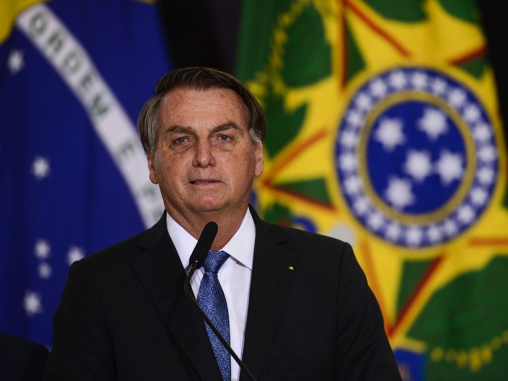 Imagem mostra Bolsonaro usando terno na frente da bandeira do Brasil e atrás de microfone