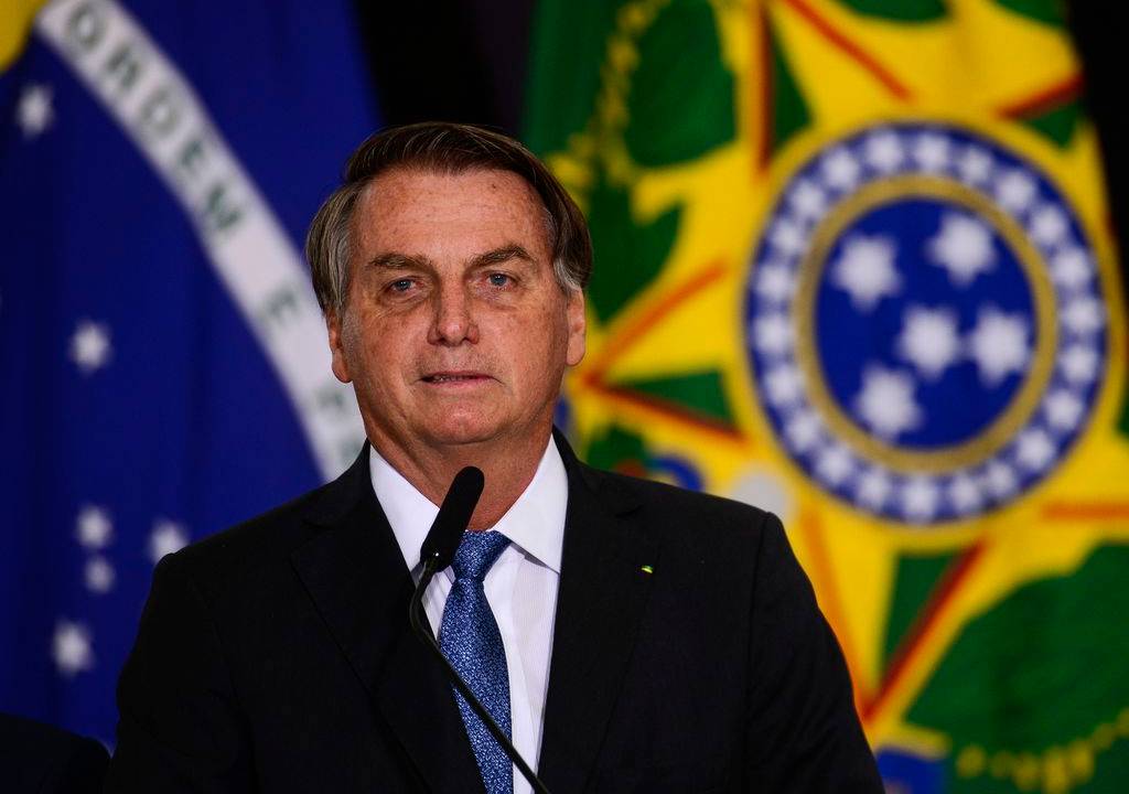 Imagem mostra Bolsonaro usando terno na frente da bandeira do Brasil e atrás de microfone