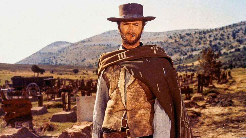 A imagem mostra Clint no Velho Oeste, vestido de cowboy, olhando para a câmera em cena de Três Homens em Conflito