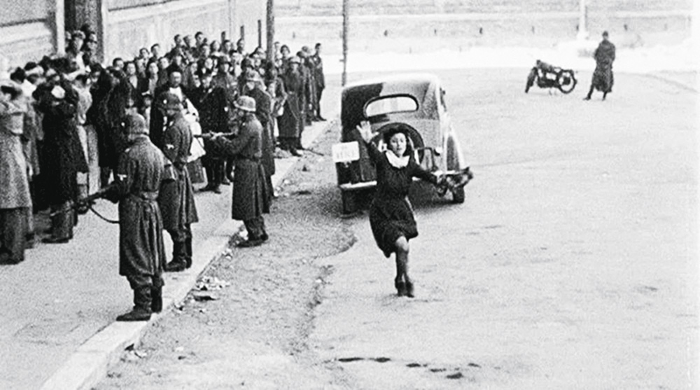 A imagem mostra uma rua cheia de pessoas, com militares e uma criança correndo no meio dela com o braço levantado