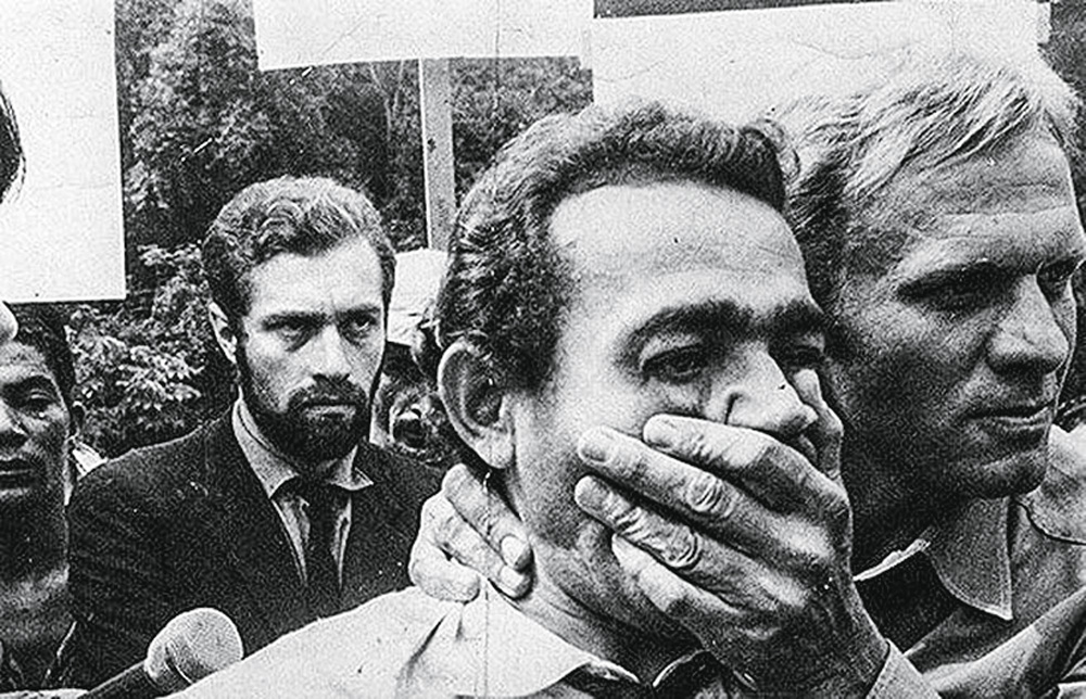 La foto mostra un uomo in mezzo alla folla, un altro uomo al suo fianco che si mette una mano sulla bocca, come se cercasse di coprirlo.