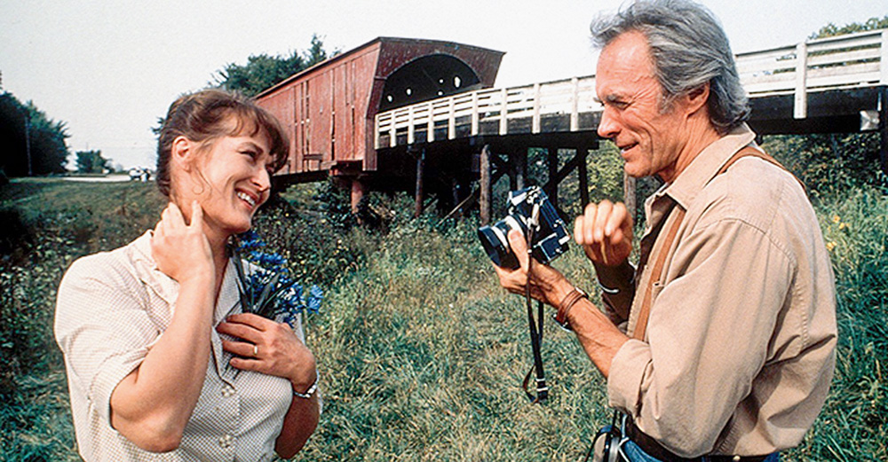 A imagem mostra Meryl e Eastwood, ambos rindo um para outro em uma região com muita vegetação próxima a uma estação de trem. Ele está com uma câmera na mãos.