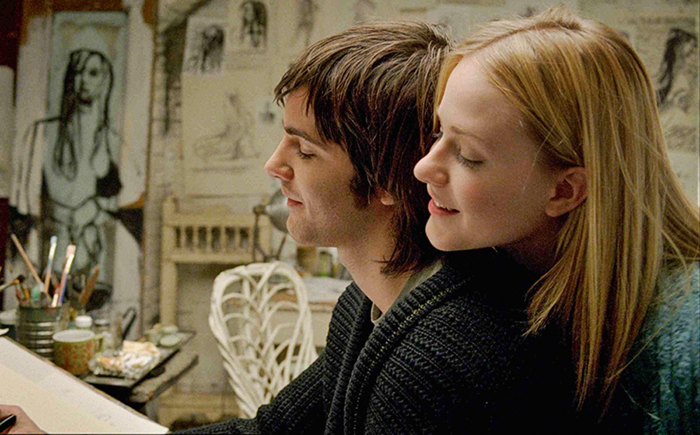 A imagem mostra o casal de Across the Universe, com uma mulher abraçando o homem por trás, ambos estão cantando, com ela com a cabeça sobre o ombro dele.