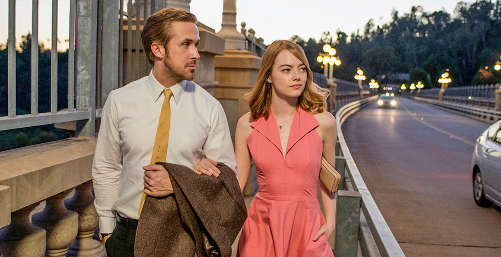 Emma Stone e Ryan Gosling aparecem na imagem em cena do filme, andando de braços entrelaçados, olhando para a rua à esquerda deles