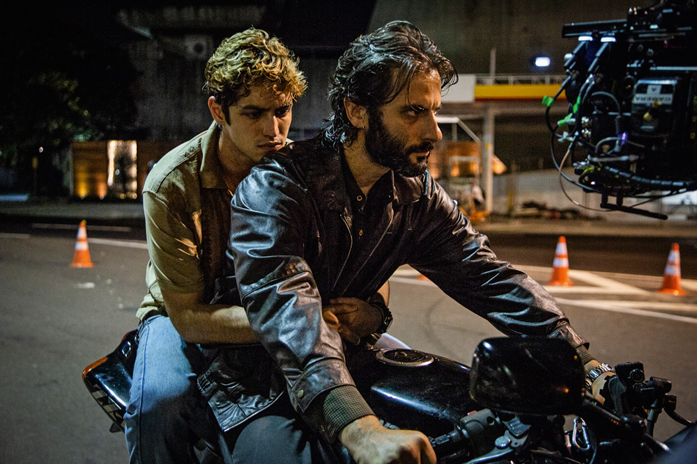 A imagem mostra os dois atores em cima de uma moto, com Gabriel abraçando Flávio, piloto do veículo.