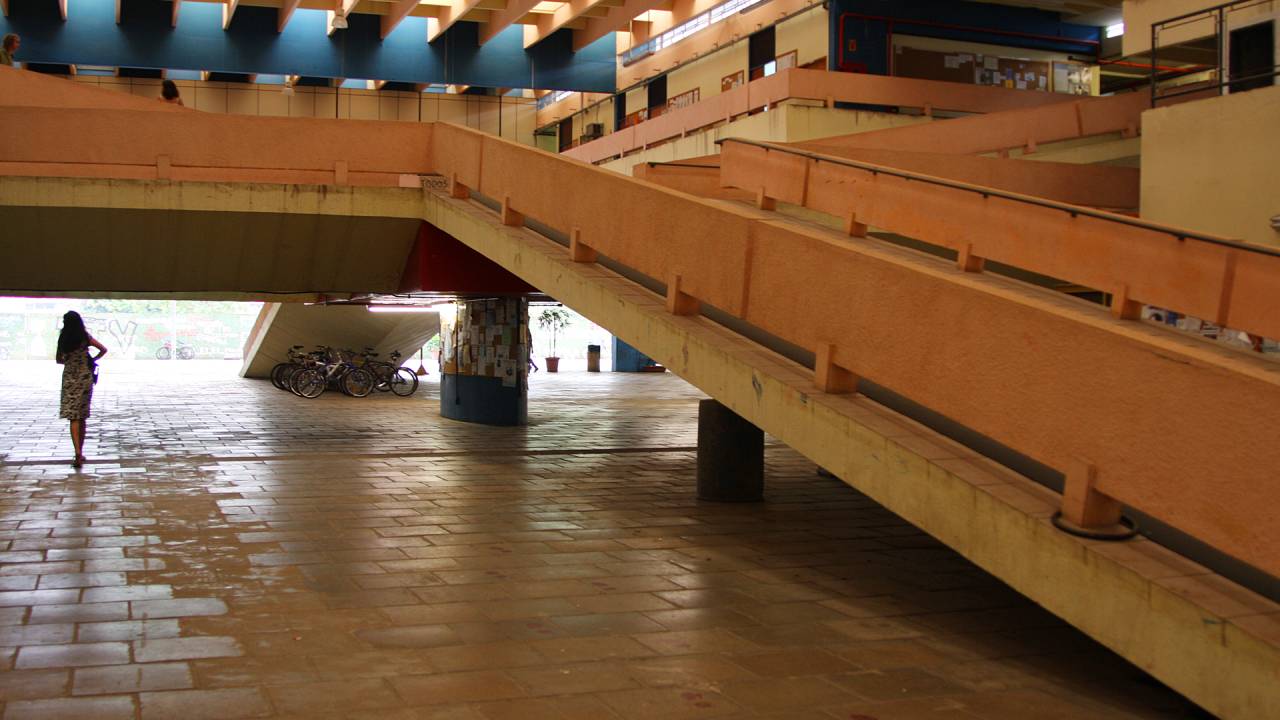 Imagem mostra foto de rampa e parte térrea de interior de prédio da USP; apenas uma pessoa aparece na imagem, uma mulher, andando, de costas