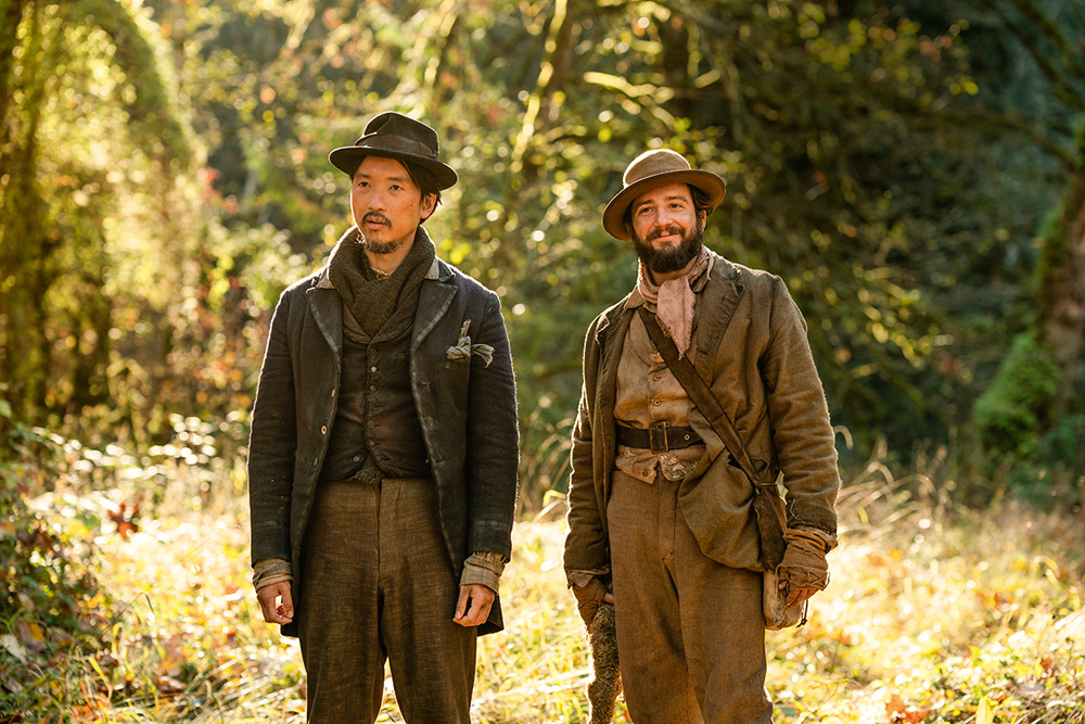 Na imagem, Orion Lee e John Magaro estão em uma floresta vestindo roupas do século XIX olhando ao seu redor