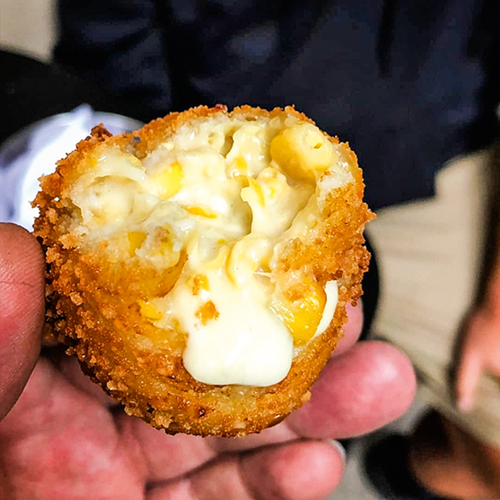 Mão segurando bolinho de milho mordido com queijo gorgonzola derretido.